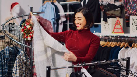 Photo for Asian customer browsing through rack of clothes in Christmas themed store, checking for fitting garment size. Woman in xmas adorn fashion boutique during winter holiday season - Royalty Free Image