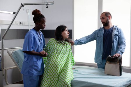 Téléchargez les photos : Infirmière obstétrique afro-américaine aidant la femme enceinte à sortir du lit dans la salle d'hôpital, préparant la patiente à la chirurgie par césarienne. Futurs parents qui attendent un enfant en maternité - en image libre de droit