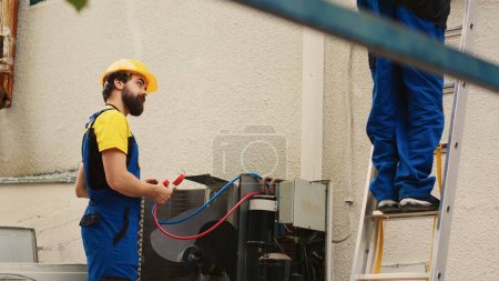 Photo for Capable electrician measuring freon levels in condenser with manifold meters while african american colleague steps down from folding ladder after finishing checking rooftop HVAC system - Royalty Free Image