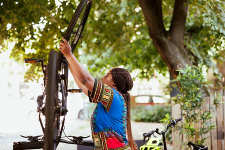 Téléchargez les photos : En été, la jeune femme active fait l'entretien annuel et l'ajustement des pneus de vélo dans la cour. Athlétique afro-américaine dame sécurise sa roue de vélo pour le cyclisme récréatif de plein air. - en image libre de droit