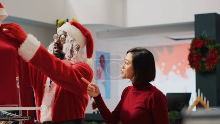 Foto de Asistente de ventas que se hace pasar por Santa Claus ayudando al cliente en la tienda de compras a encontrar la blusa roja necesaria para el traje de fiesta de Navidad. Trabajador ayudando a la mujer en la tienda de moda durante la temporada festiva - Imagen libre de derechos