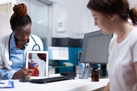 Photo for Cardiac specialist doctor explaining heart informations to patient on tablet at hospital desk. Cardiologist providing healthcare cardiac diagnosis to african american woman in clinical consultation - Royalty Free Image