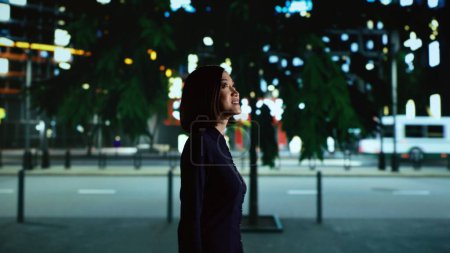 Téléchargez les photos : Asiatique se promenant près du centre-ville, regardant les gratte-ciel modernes tandis qu'elle marche sur le trottoir sous les lampadaires. Femme seule au centre-ville la nuit, souriant sur la promenade. - en image libre de droit