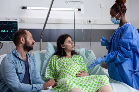 Foto de Cirujano afroamericano dando apoyo médico a la mujer embarazada, explicando la cirugía de parto en la sala de hospital. Paciente con embarazo acostada en cama con contracciones antes de entrar en trabajo de parto - Imagen libre de derechos