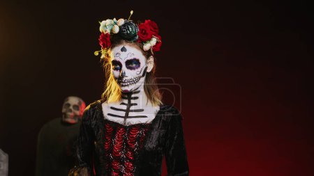 Photo for Holy goddess of death posing in halloween costume with skull make up to celebrate dios de los muertos on mexican holiday. Spooky woman as santa muerte with black and white body art. Handheld shot. - Royalty Free Image