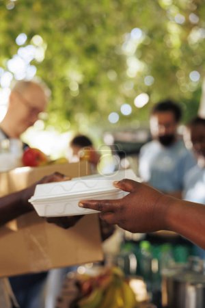 Photo for At food drive, volunteer with african american ethnicity serves a poor, hungry person a warm meal. Close-up of a less privileged needy individual receiving free food from charity worker. - Royalty Free Image