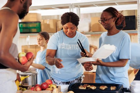 Foto de Mujeres negras amistosas que sirven comida gratis a los pobres y necesitados afroamericanos en un refugio para indigentes. Hambriento, individuo masculino menos afortunado recibe donación de comida del equipo de alivio del hambre. - Imagen libre de derechos