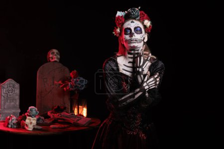 Photo for Creepy lady of death with skull make up and colorful wreath of flowers, acting like holy santa muerte on day of the dead celebration. Beautiful goddess wearing festival tradition costume. - Royalty Free Image