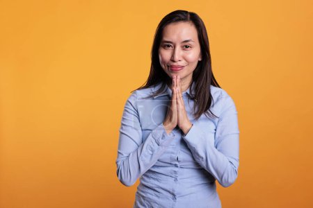 Photo for Positive asian woman praying to god in front of camera, holding hands in prayer gesture and asking for good luck. Religious model expressing hope and belief, standing in studio over yellow background - Royalty Free Image