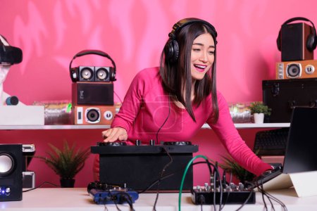 Photo for Asian performer standing at dj table mixing electronic music with techno using professional turntables, having fun in studio over pink background. Musician playing stereo sounds with electronics - Royalty Free Image