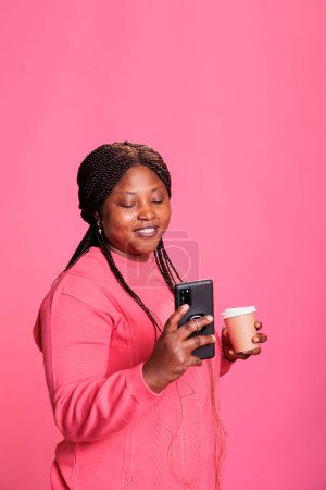 Téléchargez les photos : Femme souriante avec sweather rose tenant une tasse de café tout en lisant les nouvelles au téléphone, en postant des médias sociaux photo. Jeune adulte regarder drôle vidéo sur smartphone whle posant en studio - en image libre de droit