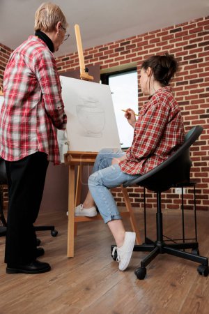 Photo for Adult daughter and senior mother doing art together while attending drawing workshop, learning to draw, developing common interests and hobbies. Teacher and student looking at canvas with artwork - Royalty Free Image