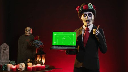 Photo for Glamorous holy entity holding laptop with greenscreen display, showing mockup template and blank copyspace in studio. Santa muerte using wireless pc with isolated chroma key screen. - Royalty Free Image