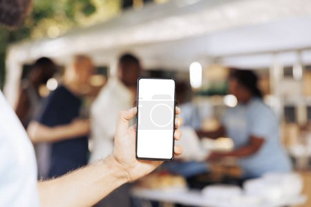 Photo for Photo focus on caucasian hand vertically grasping mobile device showing isolated mockup template at a non-profit food drive. Close-up of a person holding smartphone with white screen display. - Royalty Free Image