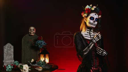 Téléchargez les photos : Flirty femme portant santa muerte body art en studio, agissant glamour le jour de la fête d'Halloween mexicaine morte. Crâne rituel art corporel, posant avec des roses noires et couronne de fleurs. - en image libre de droit