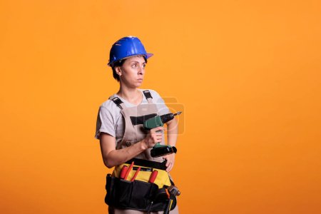 Téléchargez les photos : Ouvrière de la construction en uniforme de travail avec casque de protection tenant perceuse électrique sur fond jaune. Femme entrepreneur professionnel tenant l'équipement de travail à l'aide de la ceinture d'outils. - en image libre de droit