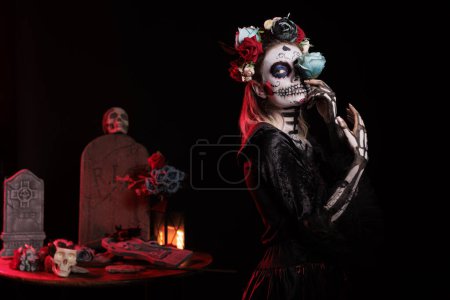 Photo for Santa muerte goddess wearing flowers crown and holding beautiful rose, looking glamour in mexican halloween costume. Celebrating dios de los muertos with tranditional festival make up. - Royalty Free Image