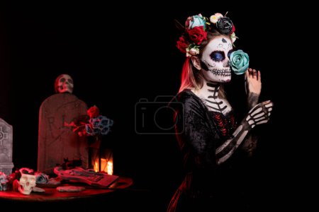 Foto de Modelo femenino con maquillaje de cráneo de azúcar y rosas azules vestidas con traje negro de muerte como santa muerte. Día del arte del cadáver para celebrar el ritual mexicano en Halloween, mito de la cultura. - Imagen libre de derechos