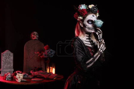 Téléchargez les photos : Femme glamour habillée en crâne de catrina en studio, portant un costume sacré traditionnel santa muerte avec maquillage noir et blanc ou art corporel. Tenir des roses et avoir une couronne de fleurs. - en image libre de droit