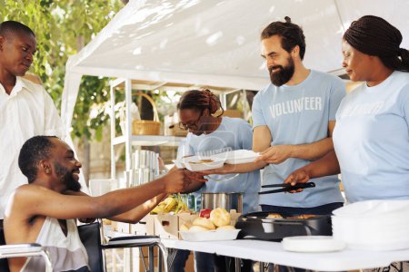 Ehrenamtliche Helfer unterschiedlicher ethnischer Zugehörigkeiten kommen mit einem Lächeln zusammen und bieten behinderten Armen und Obdachlosen Hilfe an. Wohltätigkeitsgruppe versorgte schwarzen Mann im Rollstuhl mit kostenlosem Essen.