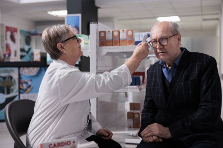 Photo for Pharmacist measuring old patient body temperature with thermometer gun, doing health check up and medical consultation. Pharmaceutical specialist testing senior man in glasses for fever in drugstore - Royalty Free Image