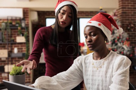Photo for Asian corporate employee helping colleague with report and pointing at laptop while working in christmas decorated office. Women in santa hats doing teamwork at new year season festive workplace - Royalty Free Image