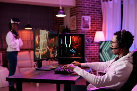 Hombre inmerso en la sesión de juego en el escritorio de la computadora, mientras que la novia en segundo plano utiliza gafas de realidad virtual. Africano americano pareja jugando juegos, divertirse en el juego de PC y vr sistema