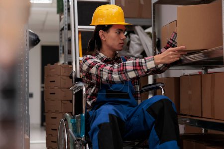 Photo for Storehouse employee in wheelchair taking cardboard box for order packing. Young asian choosing package on shelf and managing parcels transportation while working in inclusive workplace - Royalty Free Image