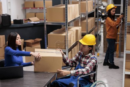 Photo for Warehouse managers handling package in post office storage room. Freight chain supply operator giving cardboard box to coworker in wheelchair while managing logistics at desk - Royalty Free Image