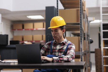 Photo for Asian warehouse delivery worker analyzing orders checklist on laptop. Young storehouse employee wearing yellow hardhat sitting at desk and checking goods supply schedule on computer - Royalty Free Image