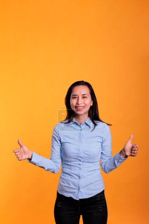 Téléchargez les photos : Femme asiatique souriante faisant le pouce vers le haut geste devant la caméra montrant l'approbation et le geste d'accord avec les doigts en studio. Modèle satisfait geste correct et symbole positif sur fond jaune - en image libre de droit