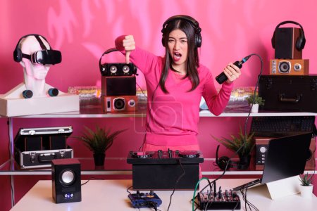 Photo for Unhappy performer holding microphone while doing thumbs down gesture, expressing disagreement in studio over pink background. Musician performing song with electronics equipment and audio instrument - Royalty Free Image