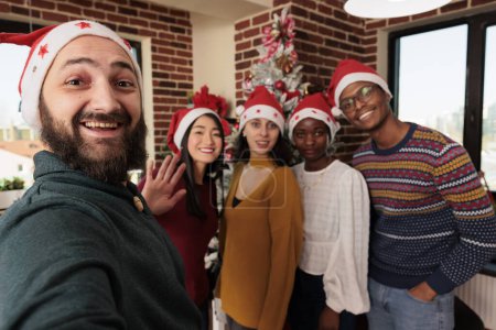 Téléchargez les photos : Équipe d'employés joyeux portant des chapeaux Santa prendre selfie tout en travaillant dans le bureau décoré de l'entreprise. Travailleurs posant pour smartphone photo tout en célébrant la veille de Noël dans l'espace de coworking - en image libre de droit