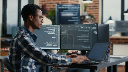 Foto de Portrait of african american developer using laptop to write code sitting at desk with multiple screens parsing algorithm in software agency. Coder working on user interface using portable computer. - Imagen libre de derechos