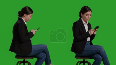 Téléchargez les photos : Close up of businesswoman browsing internet on mobile phone, acting casual on chair in studio. Company worker in suit using smartphone app to text messages, full body greenscreen backdrop. - en image libre de droit