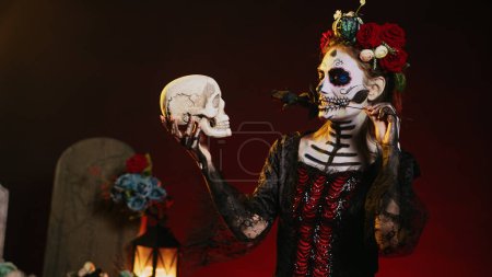 Photo for Horror female model acting flirty with skull and black roses, wearing traditional body art to celebrate mexican holiday. Looking like goddess of death on dios de los muertos ritual. Handheld shot. - Royalty Free Image