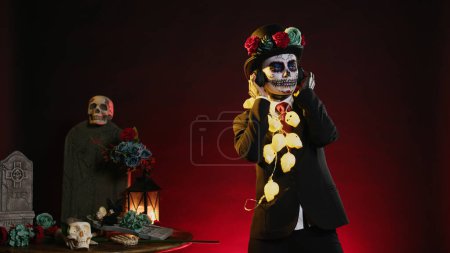 Photo for Woman in goddess of death costume dancing on music, listening to song on headphones and having fun. Wearing santa muerte skull body art to celebrate day of the dead halloween ritual. - Royalty Free Image