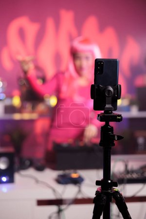 Photo for Artist with pink hair performing electronic song at turntables while filming music process with phone camera. Dj woman doing performance at nightclub with professional audio equipment - Royalty Free Image