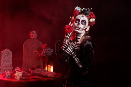 Téléchargez les photos : Dame de la mort tenant des roses pendant les vacances mexicaines, ressemblant à Santa Muerte avec costume d'Halloween et maquillage du crâne. Agissant effrayant et glamour le jour de la célébration des morts en studio. - en image libre de droit
