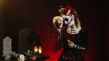 Téléchargez les photos : Femme glamour effrayant tenant crâne saint en studio, agissant effrayant et horreur pour célébrer le jour mexicain d'Halloween. Flirty déesse portant costume de festival avec art corporel, ressemblant à dame de la mort. - en image libre de droit