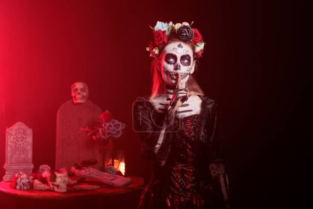 Photo for Santa muerte model showing mute confidential sign in studio, advertising hush silence symbol to keep secret. Wearing goddess of death halloween costume on traditional mexican celebration. - Royalty Free Image
