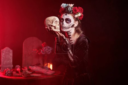 Foto de Santa muerte modelo sosteniendo cráneo en estudio, actuando de miedo y horror para celebrar halloween mexicano. Hermosa mujer en traje de festival con el arte corporal, que parece diosa de la muerte. - Imagen libre de derechos