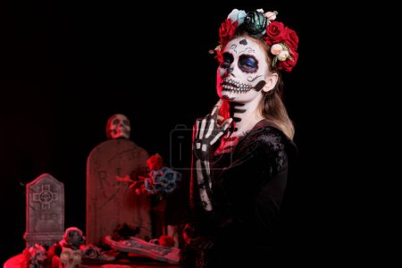 Photo for Horror lady of death wearing black glamorous costume in studio, looking like holy santa muerte with flowers crown. Acting creepy and posing dressed as goddess of dead on dios de los muertos. - Royalty Free Image