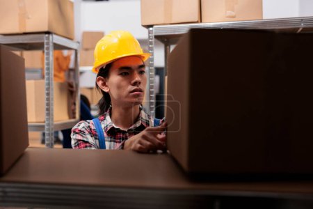 Photo for Asian young postal worker doing parcels organization in warehouse. Storehouse order picker in yellow hardhat looking at packages on shelf and searching for cardboard box close up - Royalty Free Image