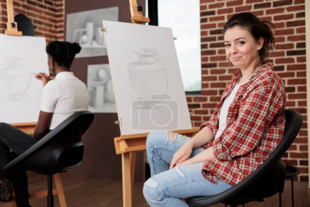 Photo for Happy young woman sitting at easel in classroom and smiling at camera, enjoying fun drawing class with friends, learning sketching techniques at group art class. Enjoyable creative hobby - Royalty Free Image