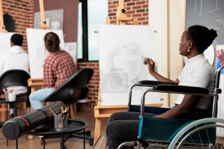 Foto de Niña afroamericana discapacitada en silla de ruedas dibujando sobre lienzo durante la clase de arte en grupo, mejorando la salud mental a través de la creatividad. Persona con discapacidad que aprende a dibujar disfrutando del proceso creativo - Imagen libre de derechos