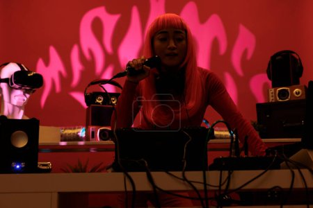 Cheeful artist with pink hair standing at dj table playing electronic music with turntables, enjoying to perform at night in club. Asian performer having fun with fans during techno concert