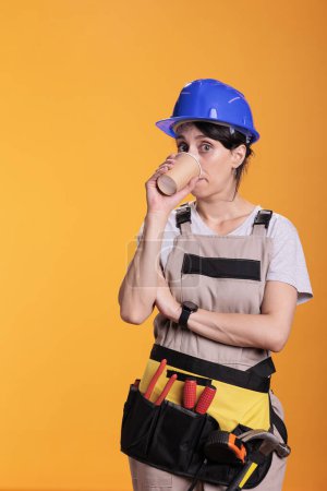 Photo for Female construction worker enjoying hot coffee during break time at work. Professional woman renovator wearing belt with tools and protective helmet in studio shot against yellow background. - Royalty Free Image