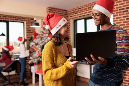 Photo for Smiling diverse colleagues laughing while doing teamwork on laptop in workplace at christmas eve. Man and woman employees in santa hats holding portable computer in decorated office - Royalty Free Image