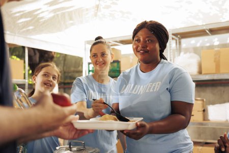Image showcasing friendly multicultural women serving free, hot meals to homeless caucasian individual at a food drive. Volunteer crew helping and feeding the hungry and poor at an outdoor center.